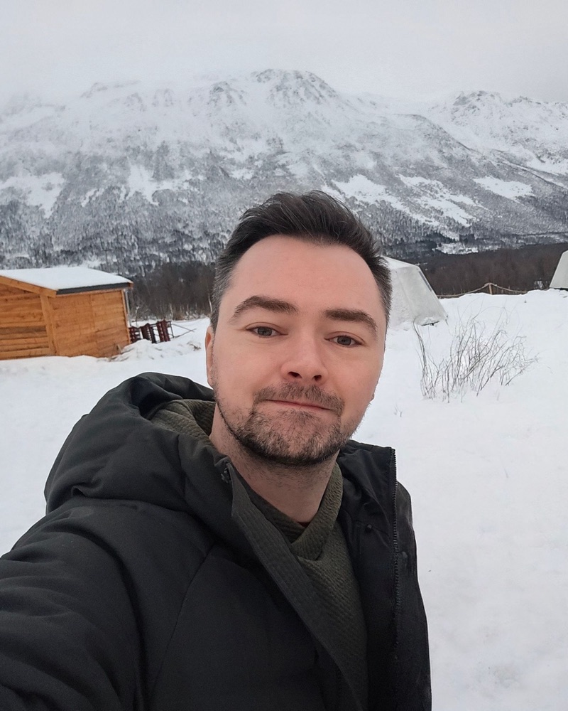 Adam Pritchard in a winter coat, standing in the snow with a mountain behind him